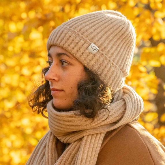 A woman wears a beige scarf and hat made of fine camel wool in front of yellow autumn leaves