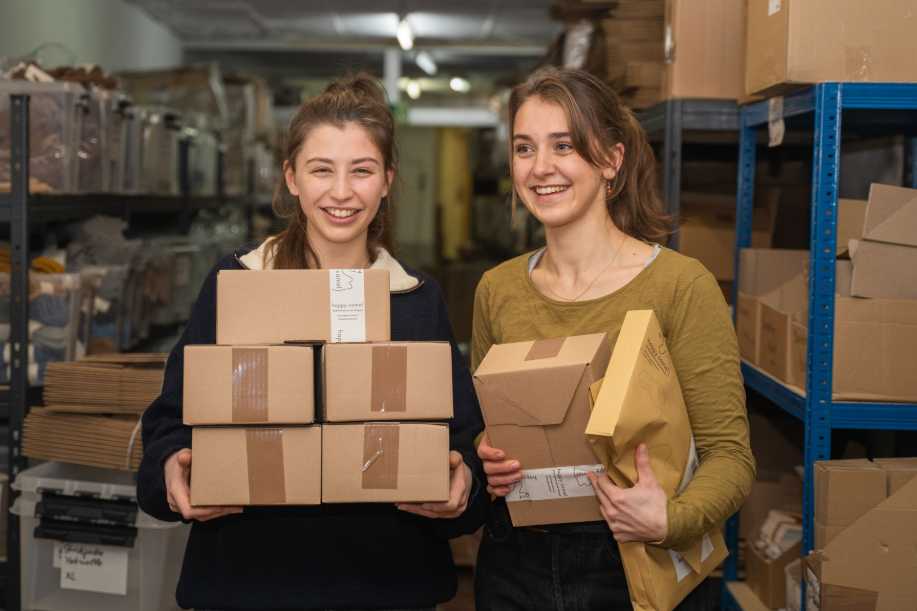 Zwei junge Frauen lachen mit Paketen im Arm in die Kamera in einem Lagerkeller