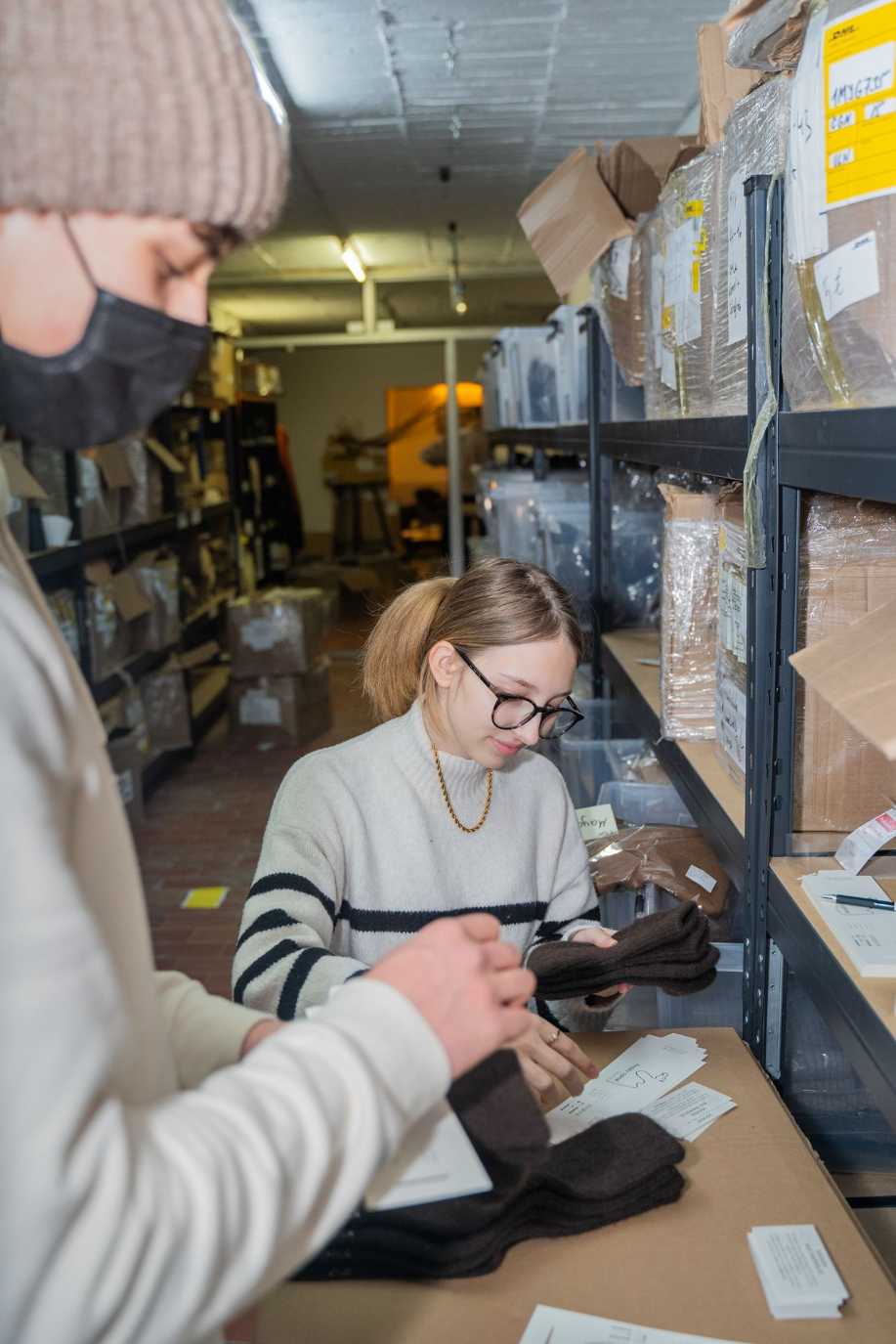 Eine Frau in weißem Pullover packt Kisten zwischen zwei Regalen