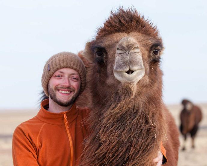 Malte 2016 in der Wüste Gobi Ein junger Mann mit Mütze steht neben einem Kamel und beide schauen in die Kamera.