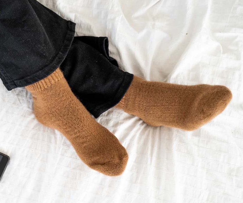 Socken aus Kamelwolle, braun Füße in braunen Socken überschlagen auf einem weißen Bett